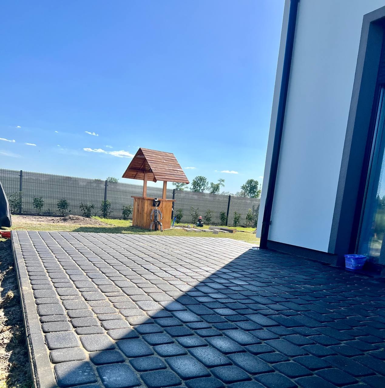 Ułożona, ciemnoszara kostka brukowa na posesji z widoczną studnią ogrodową z drewnianą obudową i nowoczesnym domem w tle, oświetlona jasnym słońcem.