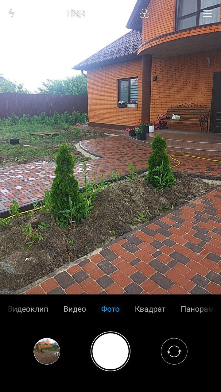 Ułożona kostka brukowa w dwóch odcieniach czerwieni i szarości, prowadząca do wejścia do domu z cegły; świeżo posadzone małe drzewka iglaste wzdłuż ścieżki.