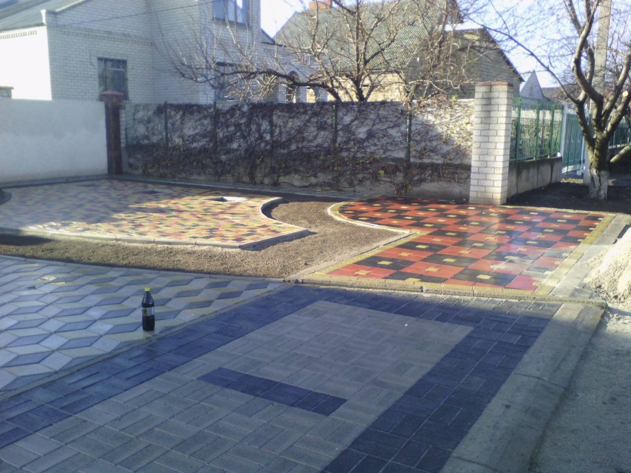 Ułożona nawierzchnia z kostki brukowej w kilku wzorach i kolorach, z butelką napoju w rogu, na tle ogrodzenia i budynku mieszkalnego.