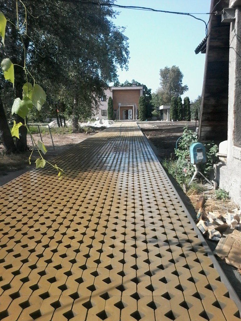 Długa, nowo ułożona ścieżka z beżowej kostki brukowej o geometrycznym wzorze, prowadząca do budynku w oddali, z widocznym drzewem po lewej stronie i elementami budowlanymi po prawej.