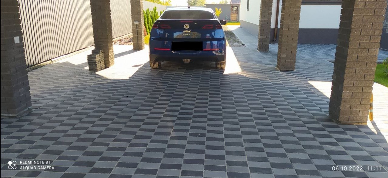 Dwukolorowa kostka brukowa ułożona w szachownicę pod zadaszeniem, z zaparkowanym niebieskim samochodem w tle.