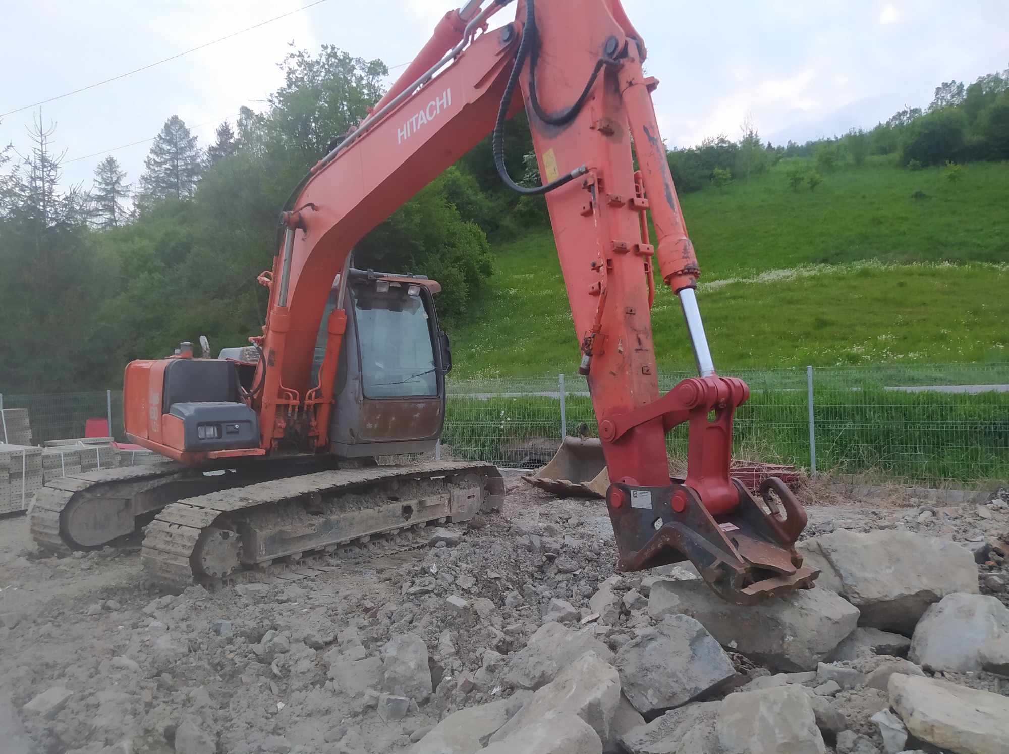 Pomarańczowa koparka Hitachi na nierównym terenie z dużymi kamieniami, w tle zielone wzgórza i drzewa.