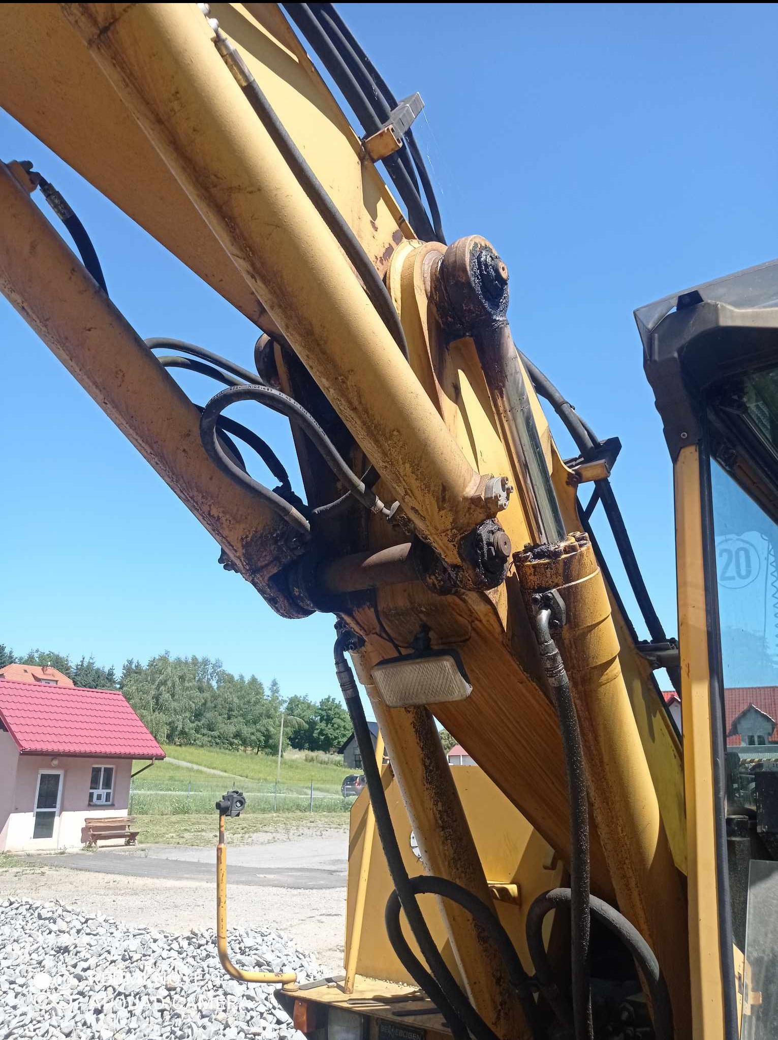 Żółte ramię koparki hydraulicznej z widocznymi przewodami i elementami hydraulicznymi, zarys kabiny operatora po prawej stronie, w tle budynek z czerwonym dachem i błękitne niebo.