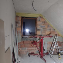 Paweł Dworakowski - Wnętrze poddasza w trakcie adaptacji: ściany częściowo obłożone płytami gipsowo-kartonowymi, widoczny podnośnik do płyt, drabina aluminiowa i narzędzia, surowa cegła wokół okna.