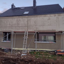 Paweł Dworakowski - Dom w trakcie tynkowania elewacji zewnętrznej, widoczne rusztowania, drabina oparta o ścianę, ściana pokryta szarym tynkiem, dach z ciemnej dachówki, okna dachowe.