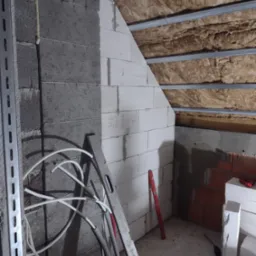 Wnętrze poddasza w trakcie prac budowlanych: murowana ściana z betonu komórkowego, izolacja wełną mineralną pod metalową konstrukcją dachu, poziomica oparta o ścianę, przewody elektryczne.