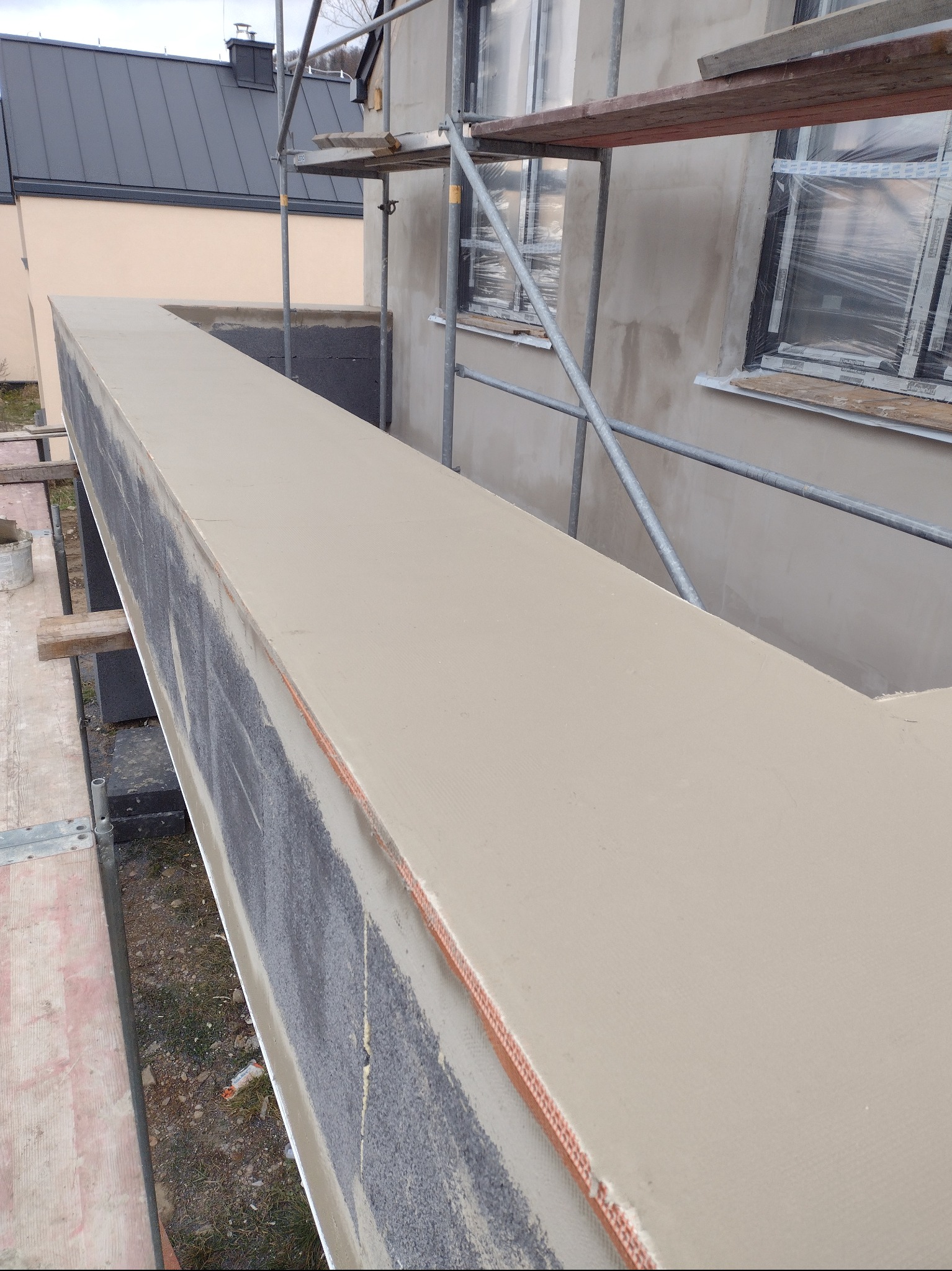 Świeżo otynkowana elewacja balkonu z widocznymi rusztowaniami i zabezpieczonymi oknami folią.