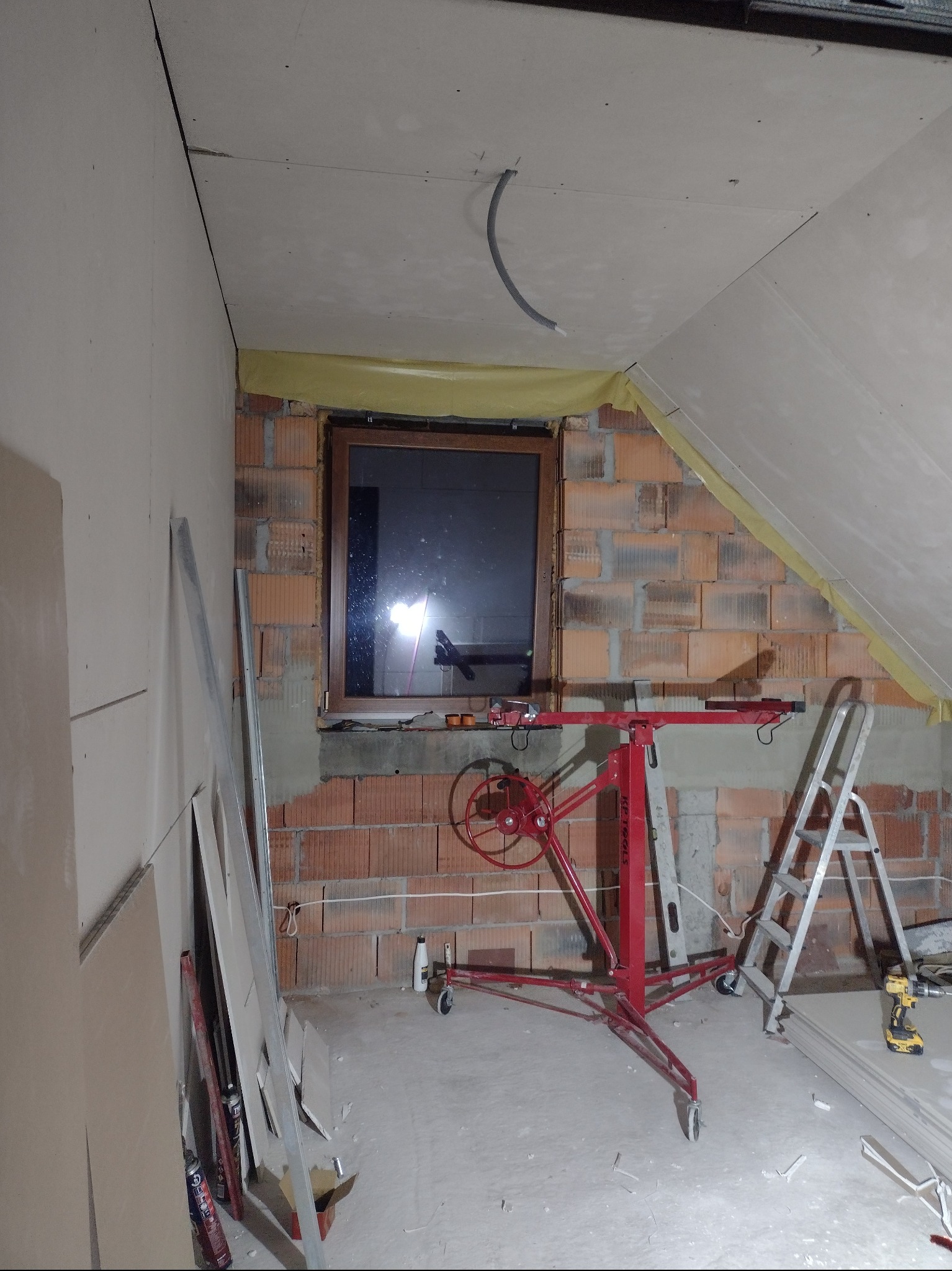 Wnętrze poddasza w trakcie adaptacji: ściany częściowo obłożone płytami gipsowo-kartonowymi, widoczny podnośnik do płyt, drabina aluminiowa i narzędzia, surowa cegła wokół okna.