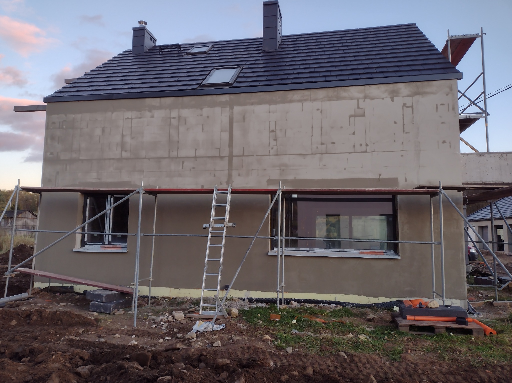 Dom w trakcie tynkowania elewacji zewnętrznej, widoczne rusztowania, drabina oparta o ścianę, ściana pokryta szarym tynkiem, dach z ciemnej dachówki, okna dachowe.