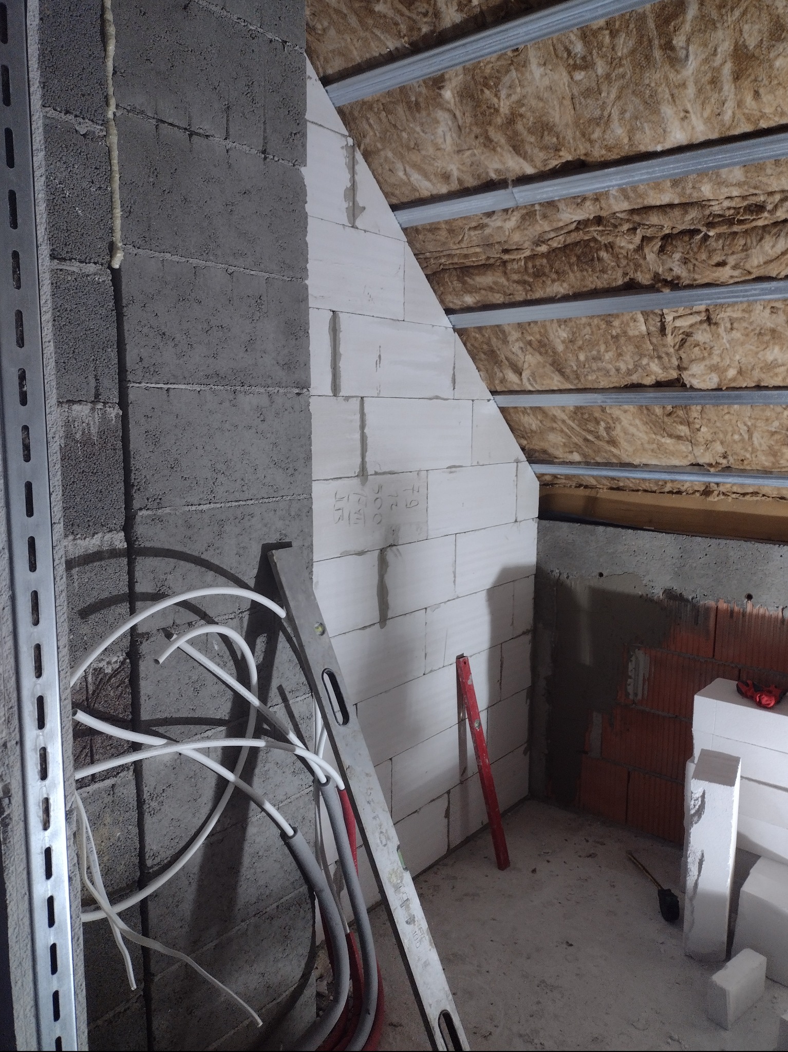 Wnętrze poddasza w trakcie prac budowlanych: murowana ściana z betonu komórkowego, izolacja wełną mineralną pod metalową konstrukcją dachu, poziomica oparta o ścianę, przewody elektryczne.