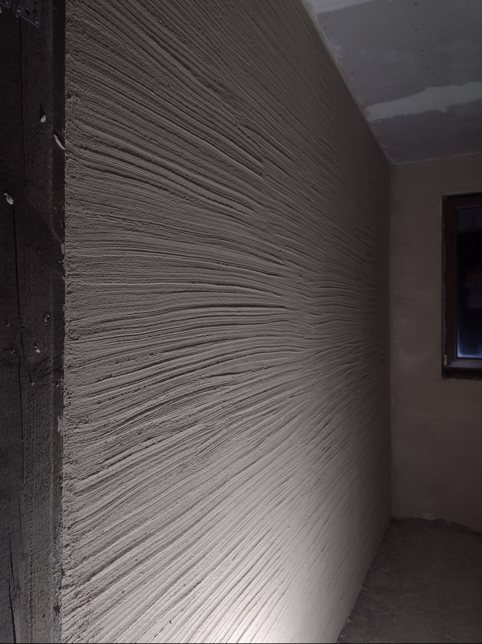 Świeżo nałożony tynk maszynowy ze specyficzną, rowkowaną fakturą, widoczny w rogu pomieszczenia z surowym, ciemnym słupem i oknem w tle.