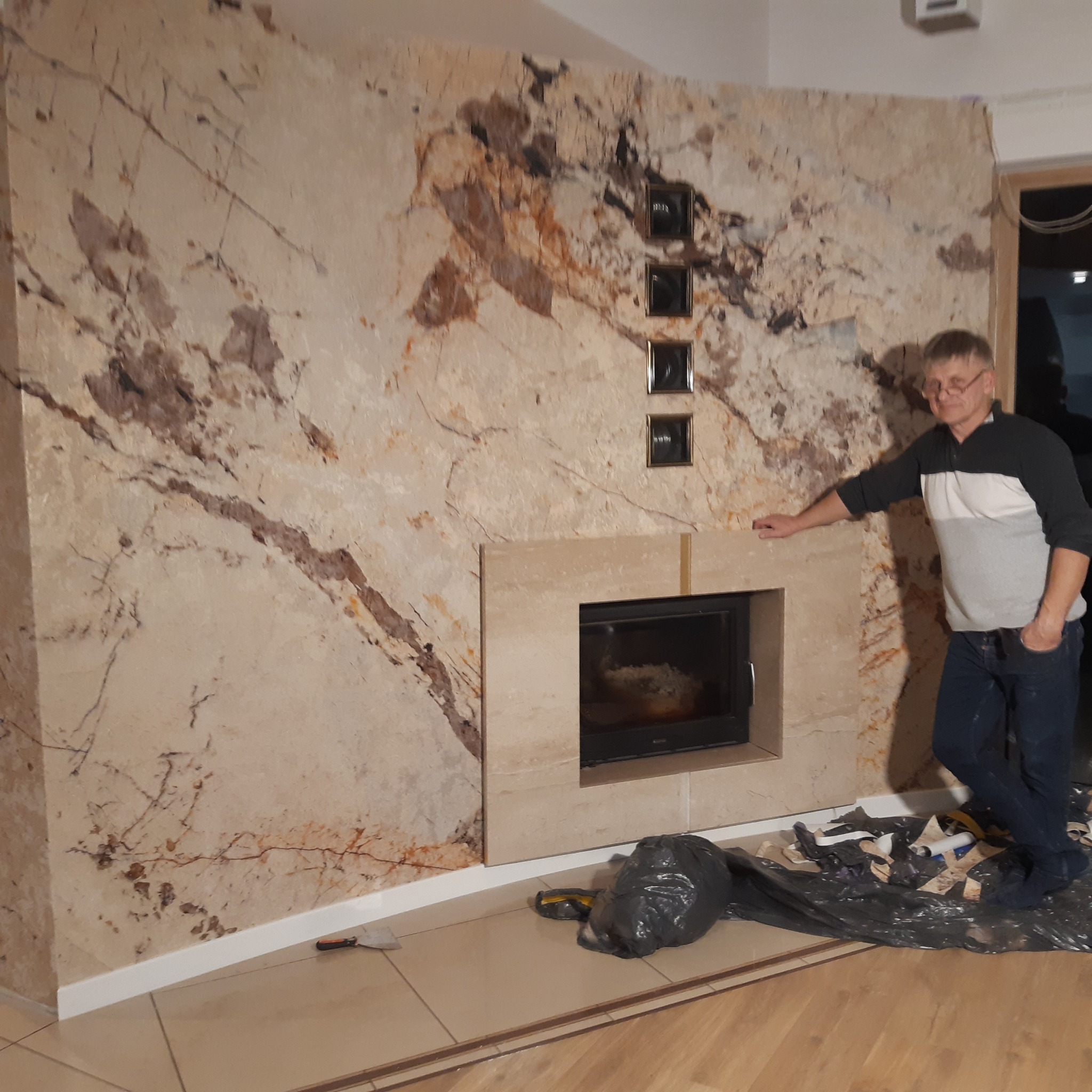 Ściana z kamienia dekoracyjnego z wbudowanym kominkiem, ozdobiona trzema małymi ramkami. Mężczyzna opiera się o kominek. Na podłodze narzędzia i worek na śmieci.