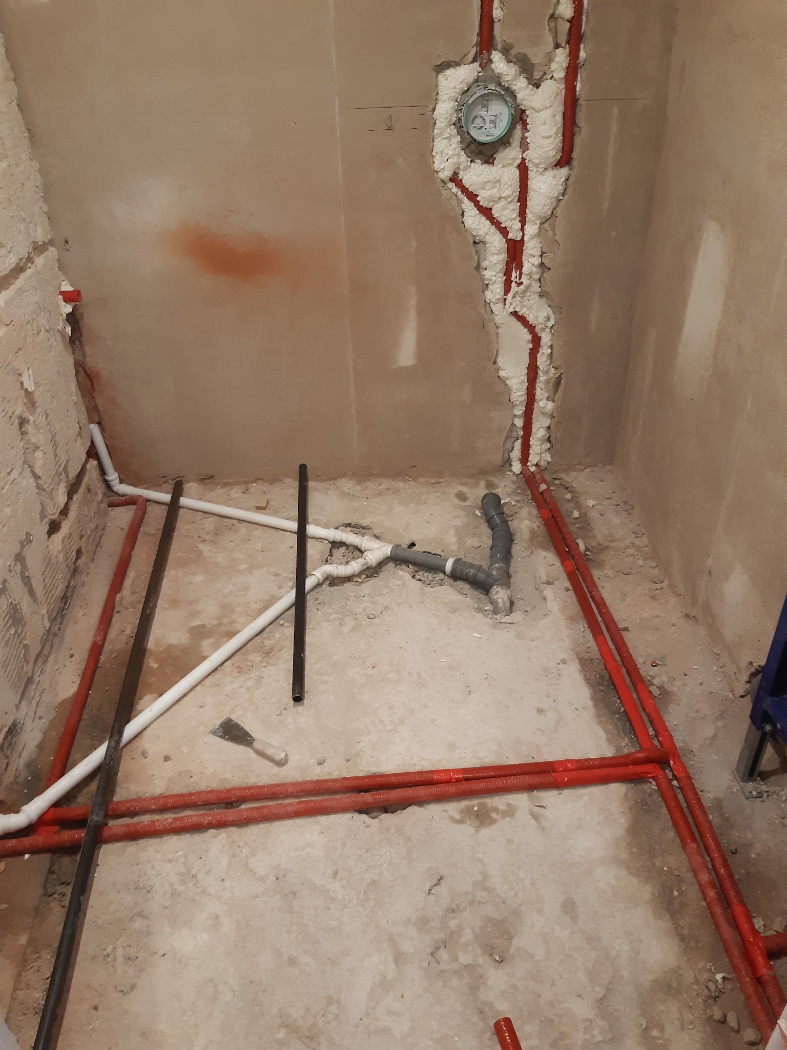 Instalacja wodno-kanalizacyjna w trakcie remontu łazienki: czerwone rury w ścianie zabezpieczone pianką montażową, białe i szare rury na podłodze, widoczna szpachelka.