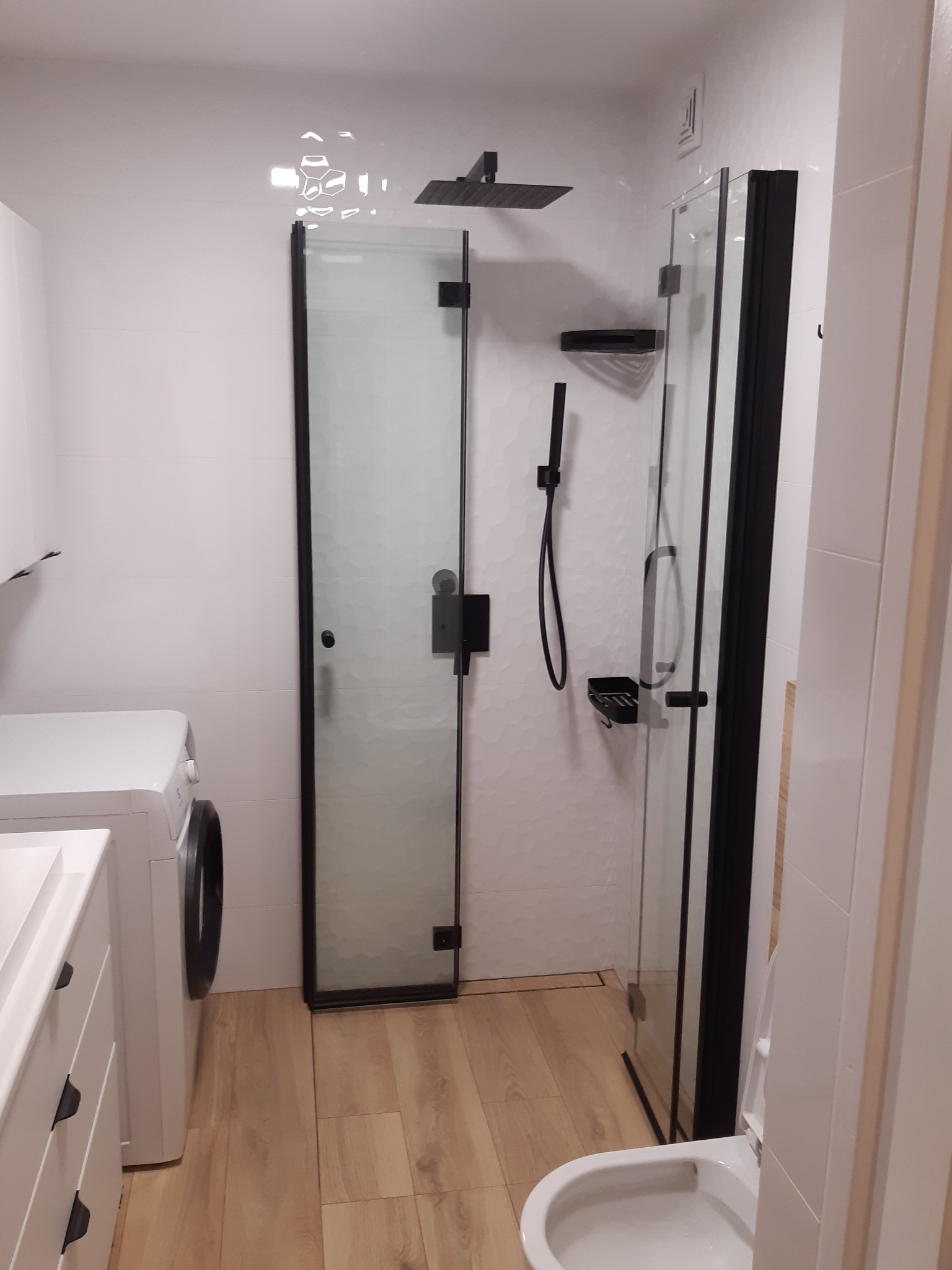 Nowoczesna łazienka z kabiną prysznicową typu walk-in z czarnymi profilami, białe płytki heksagonalne na ścianie, drewnopodobne panele na podłodze i biała pralka.