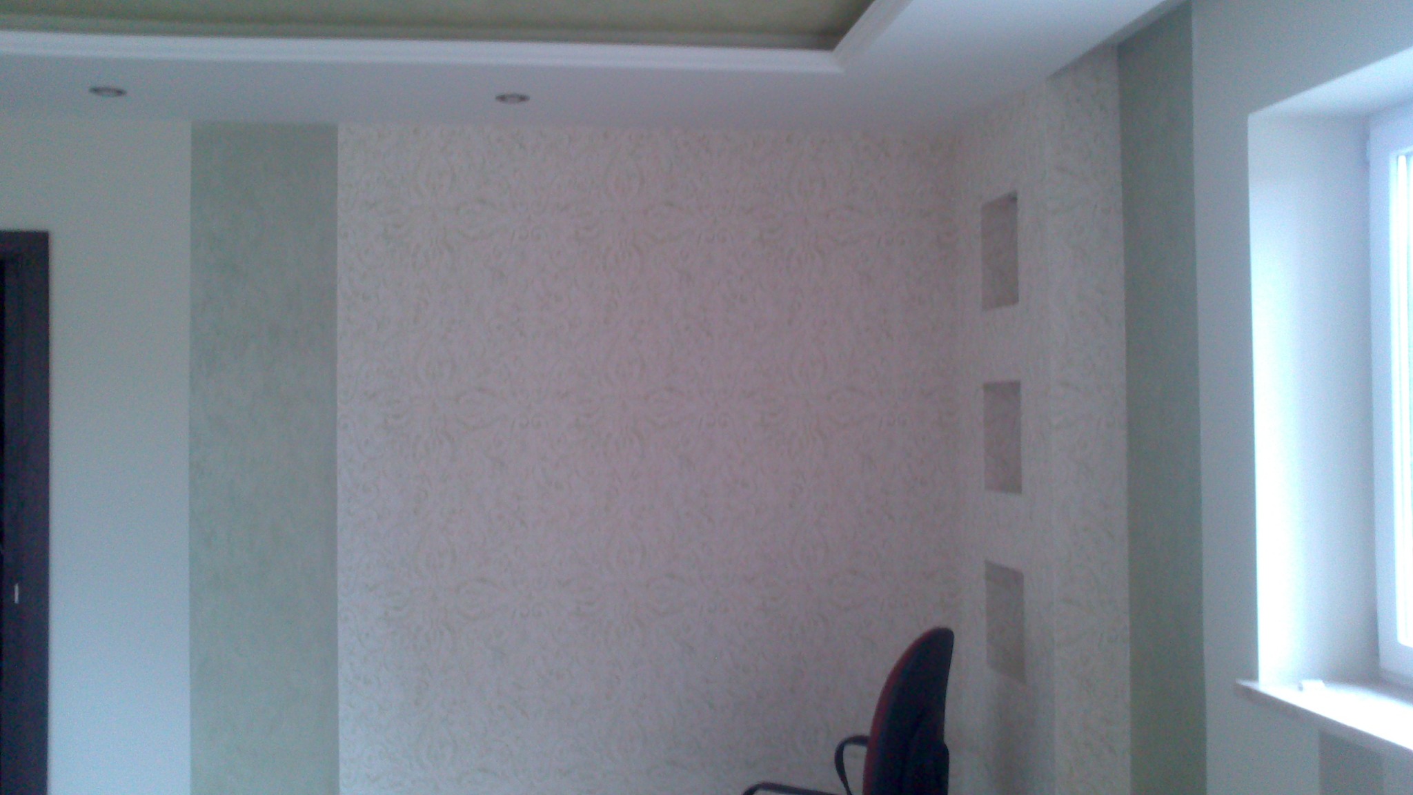 Wnętrze pokoju po tapetowaniu z wzorzystą tapetą i pomalowanymi na szaro kolumnami. Widoczne wnęki w ścianie i fragment krzesła biurowego.