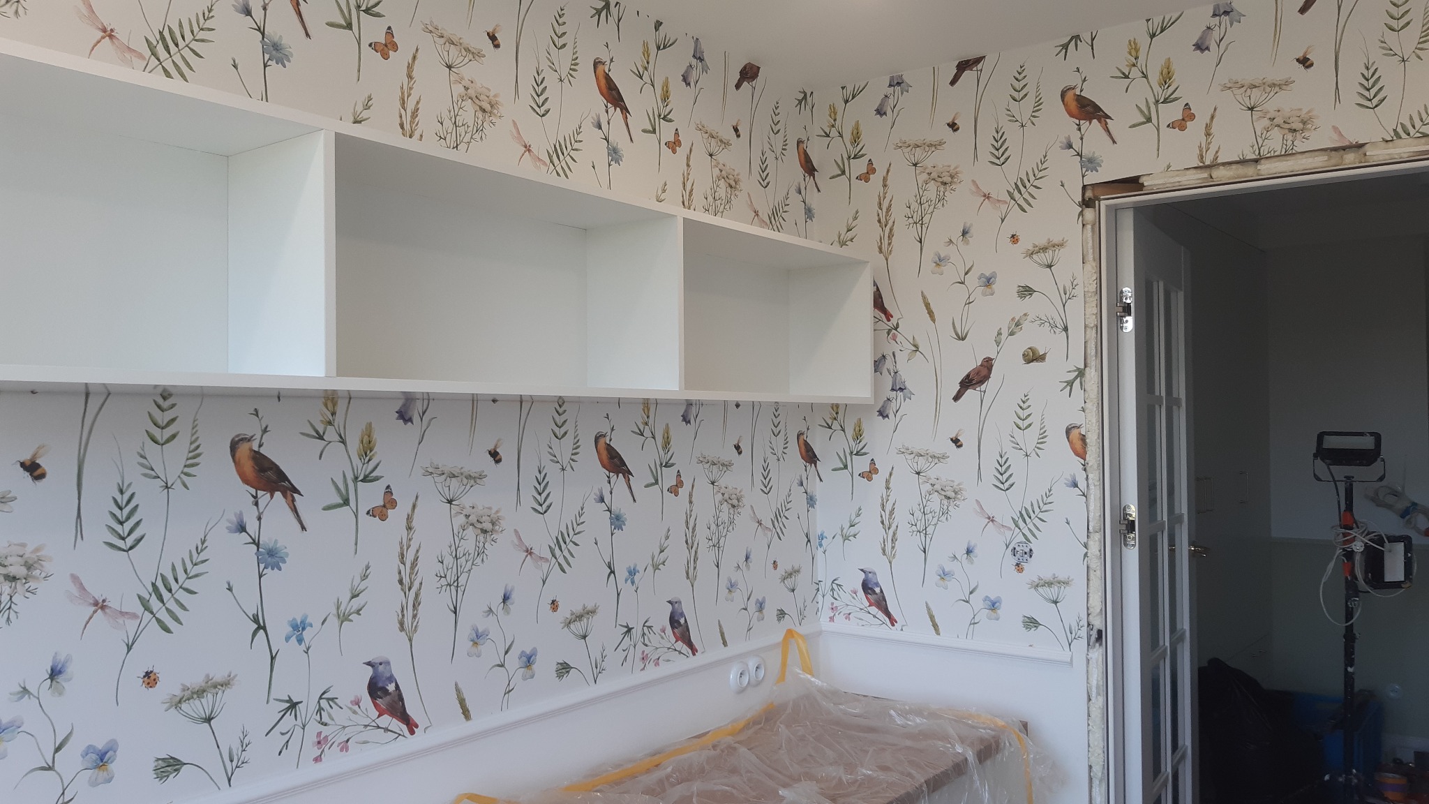Pokój po tapetowaniu z motywem roślinnym i ptaków. Białe półki wiszące i listwa przypodłogowa. Widoczne przygotowania do montażu drzwi.