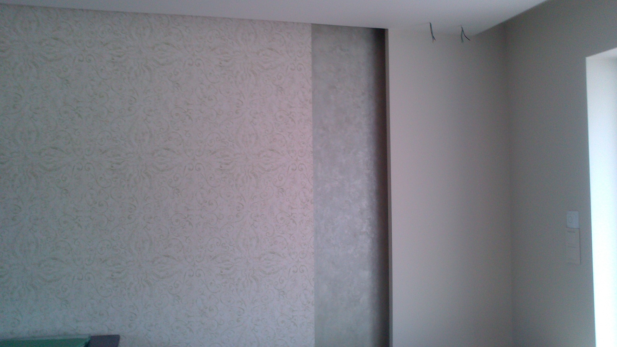 Ściana po tapetowaniu z delikatnym wzorem i fragmentem pomalowanym na szaro, widoczne wystające kable ze ściany, jasne wnętrze.