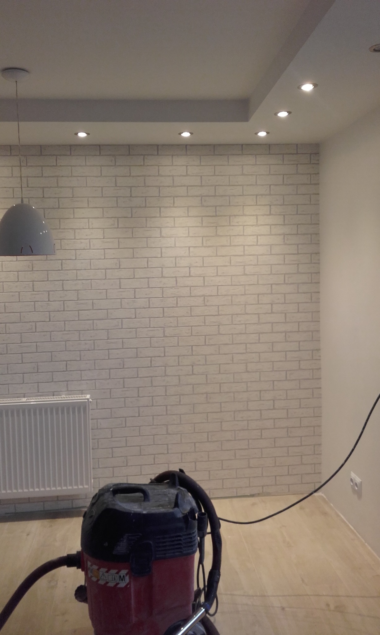 Ściana wykończona białą cegłą dekoracyjną, oświetlona punktowo, z widocznym grzejnikiem i odkurzaczem przemysłowym. Nowoczesne wykończenie wnętrza.