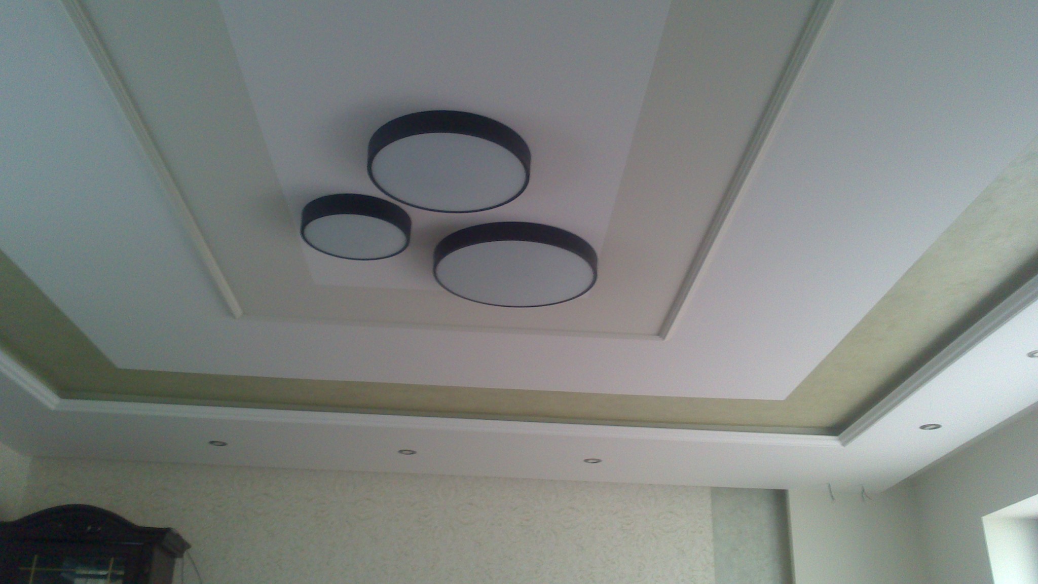 Dekoracyjny sufit podwieszany z oświetleniem punktowym i trzema okrągłymi lampami. Kremowe i białe wykończenie, wzorzysta tapeta w tle.