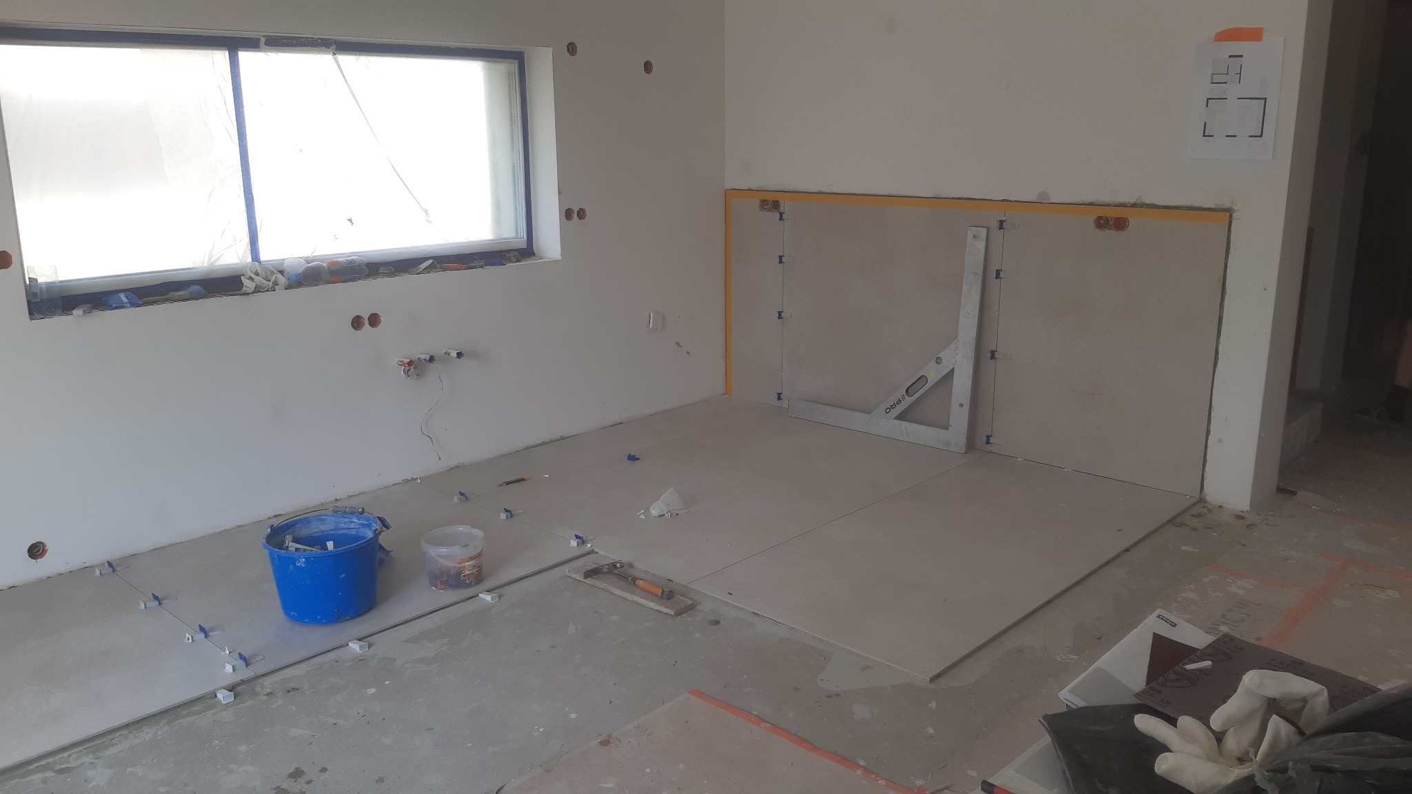 Wnętrze w trakcie prac glazurniczych: ułożone płytki podłogowe, poziomica przy ścianie, niebieskie wiadro, okno zabezpieczone folią, widoczne narzędzia i akcesoria glazurnicze.