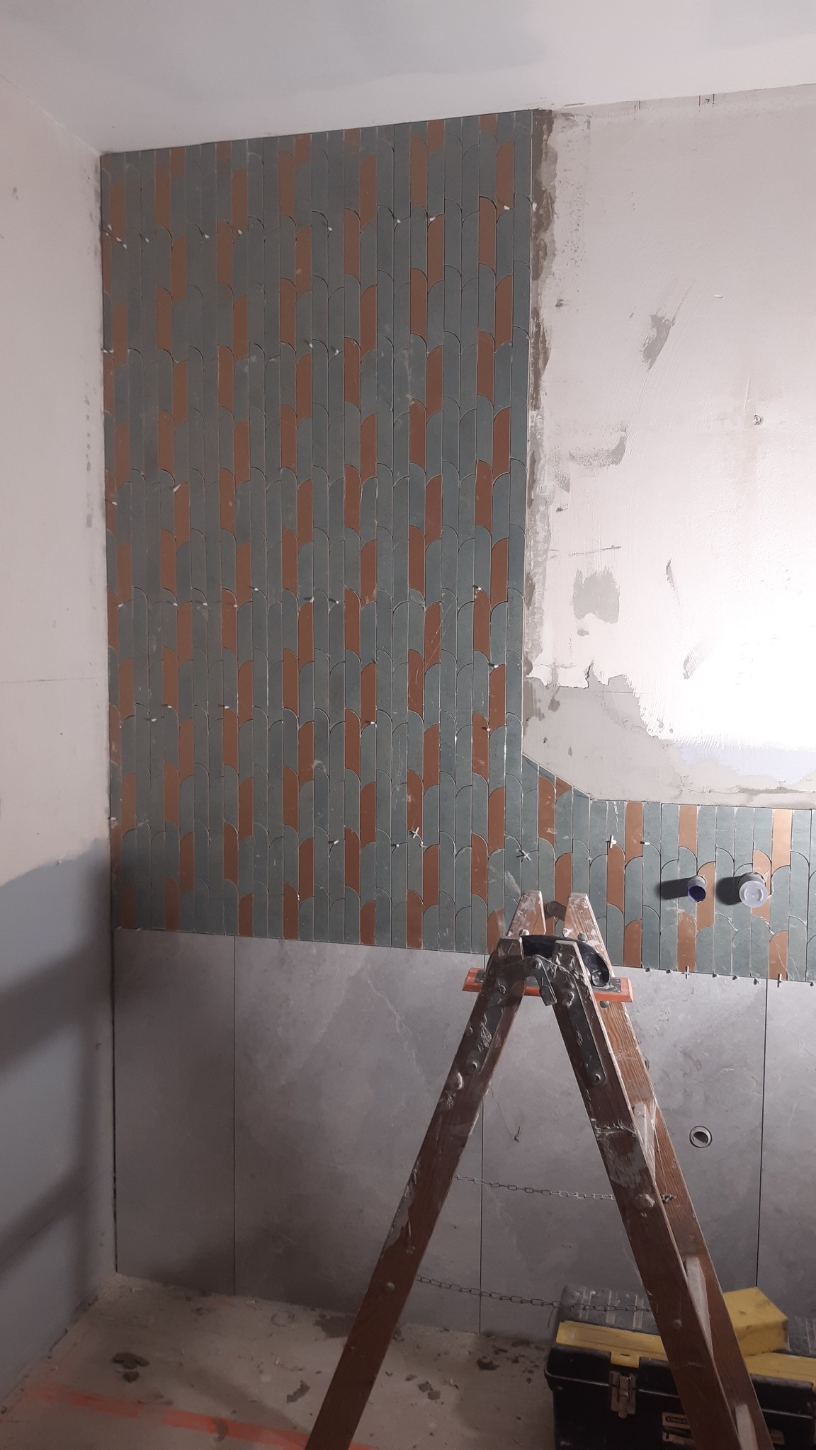 Fragment ściany łazienki z ułożonymi, pionowymi płytkami w odcieniach szarości i miedzi, częściowo wykończonej. Na pierwszym planie drewniana drabina i skrzynka narzędziowa.