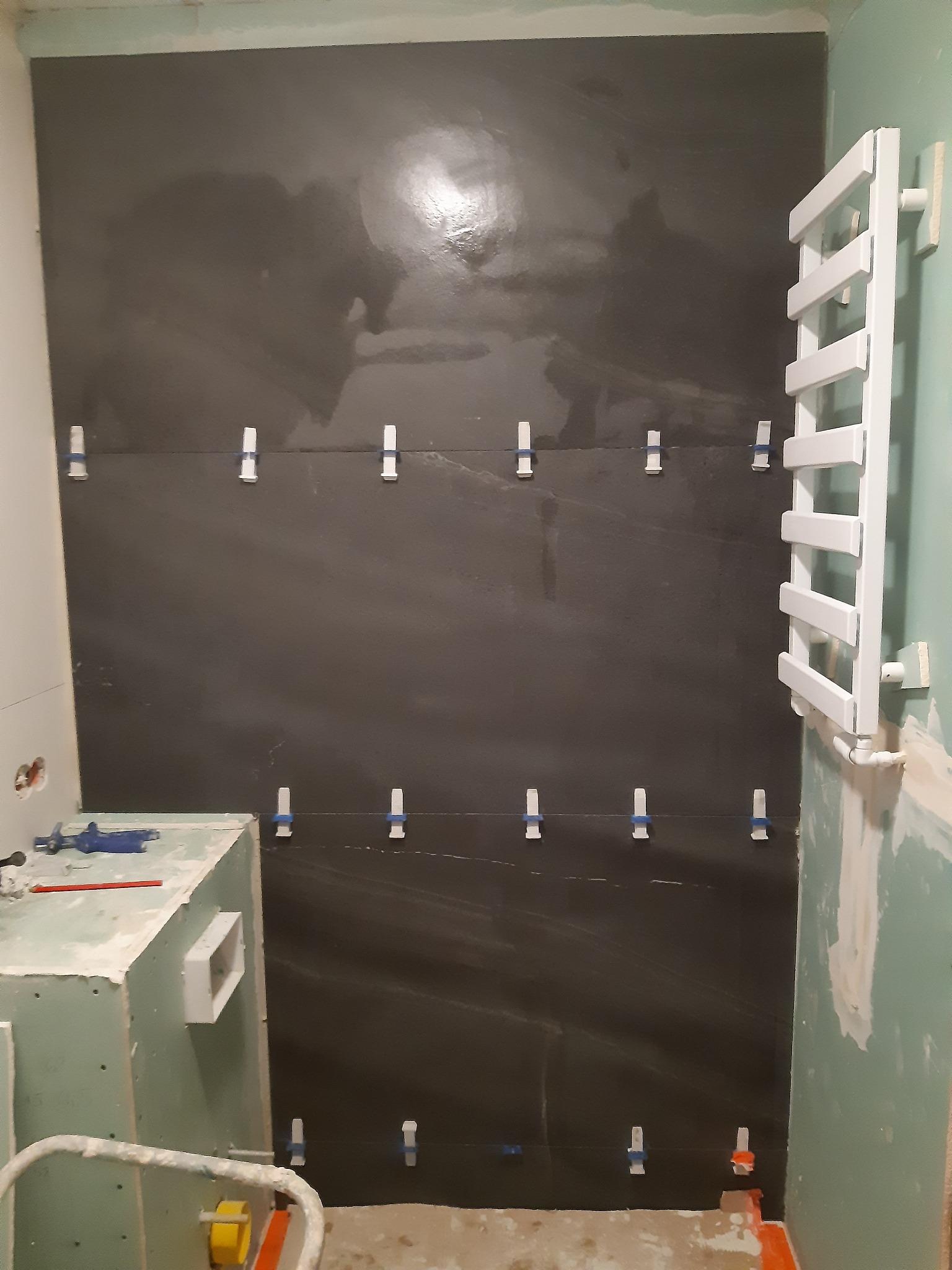 Ciemne płytki na ścianie z białymi krzyżykami dystansowymi w trakcie układania. Grzejnik drabinkowy w tle. Prace glazurnicze w Brzegu.