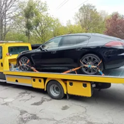 Czarna Panamera zabezpieczona pomarańczowymi pasami transportowymi na żółtej lawecie z włączonymi światłami ostrzegawczymi na dachu, widok z boku na tle drzew.