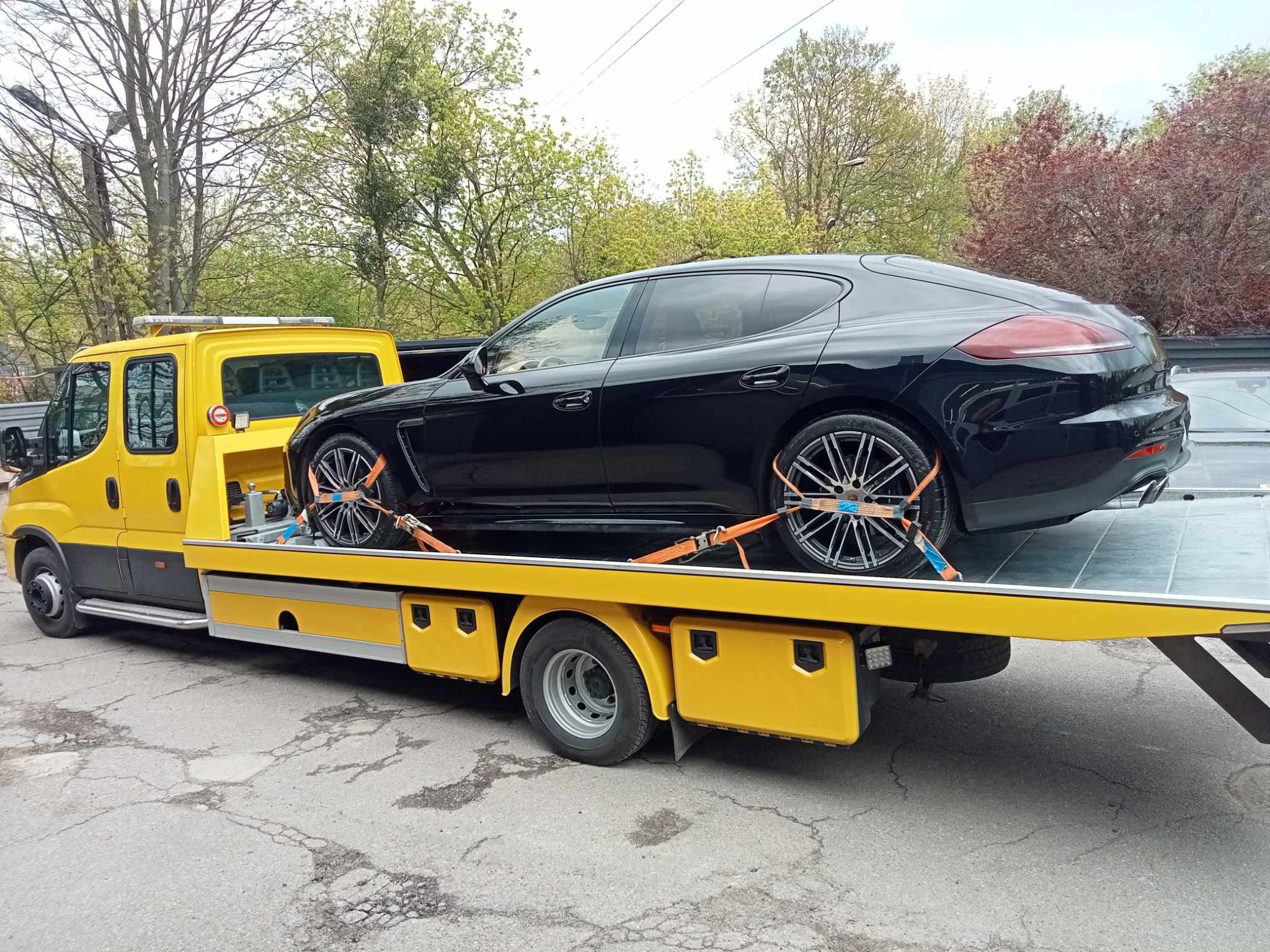 Czarna Panamera zabezpieczona pomarańczowymi pasami transportowymi na żółtej lawecie z włączonymi światłami ostrzegawczymi na dachu, widok z boku na tle drzew.