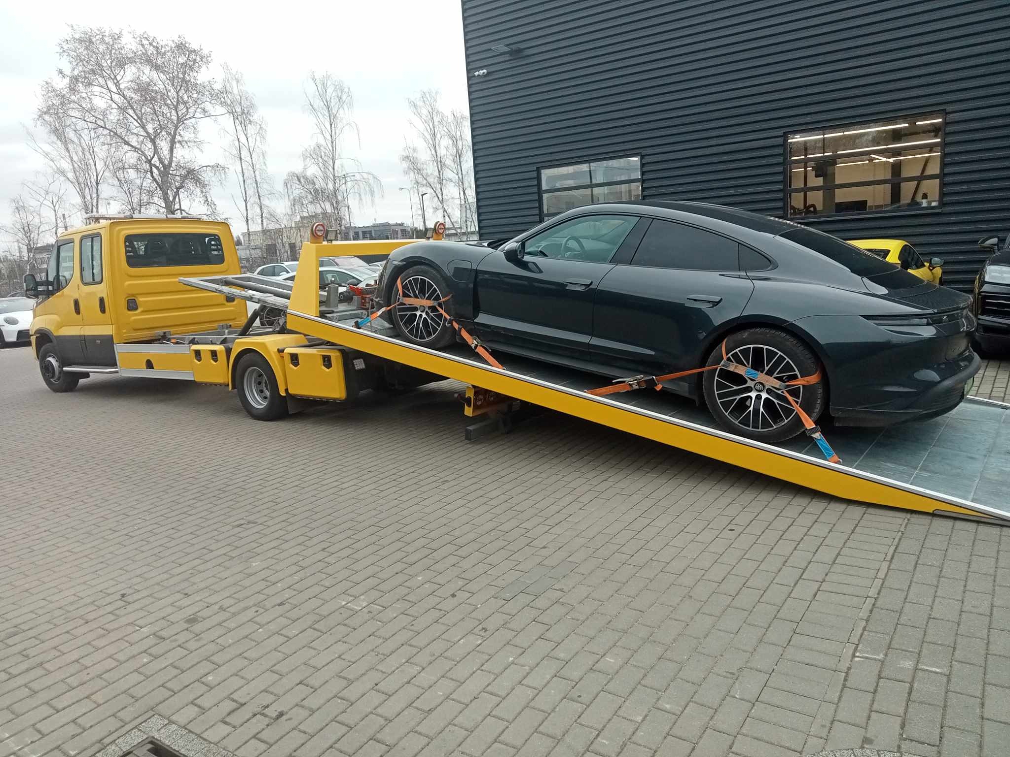 Żółty pojazd transportowy z platformą załadunkową przewozi ciemnozielony samochód osobowy zabezpieczony pasami transportowymi na tle nowoczesnego budynku z ciemną elewacją.