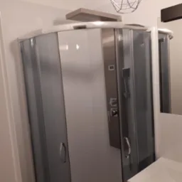 Kabina prysznicowa z szarymi szybami i panelem natryskowym w wykończonej łazience z białymi płytkami, oświetlona nowoczesną lampą wiszącą.