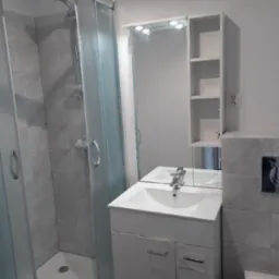 Nowoczesna łazienka z kabiną prysznicową z zaokrąglonym profilem, białą szafką umywalkową z lustrem i szafką wiszącą z półkami, wykończona szarą płytką imitującą marmur.