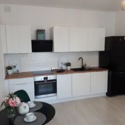 Nowoczesna, jasna kuchnia z białymi szafkami, drewnianym blatem, czarną lodówką i płytą indukcyjną, ozdobiona obrazkami i kwiatami na stole