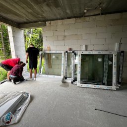 Okna Drzwi Bramy Rolety - Trzech mężczyzn przygotowuje się do montażu białych okien z profili VEKA w surowym stanie budynku z betonu komórkowego; widoczne profile okienne w folii ochronnej.