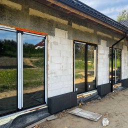 Okna Drzwi Bramy Rolety - Montaż okien z czarnymi ramami w surowym stanie domu z betonu komórkowego, widoczne pianowanie i folia hydroizolacyjna przy fundamentach.