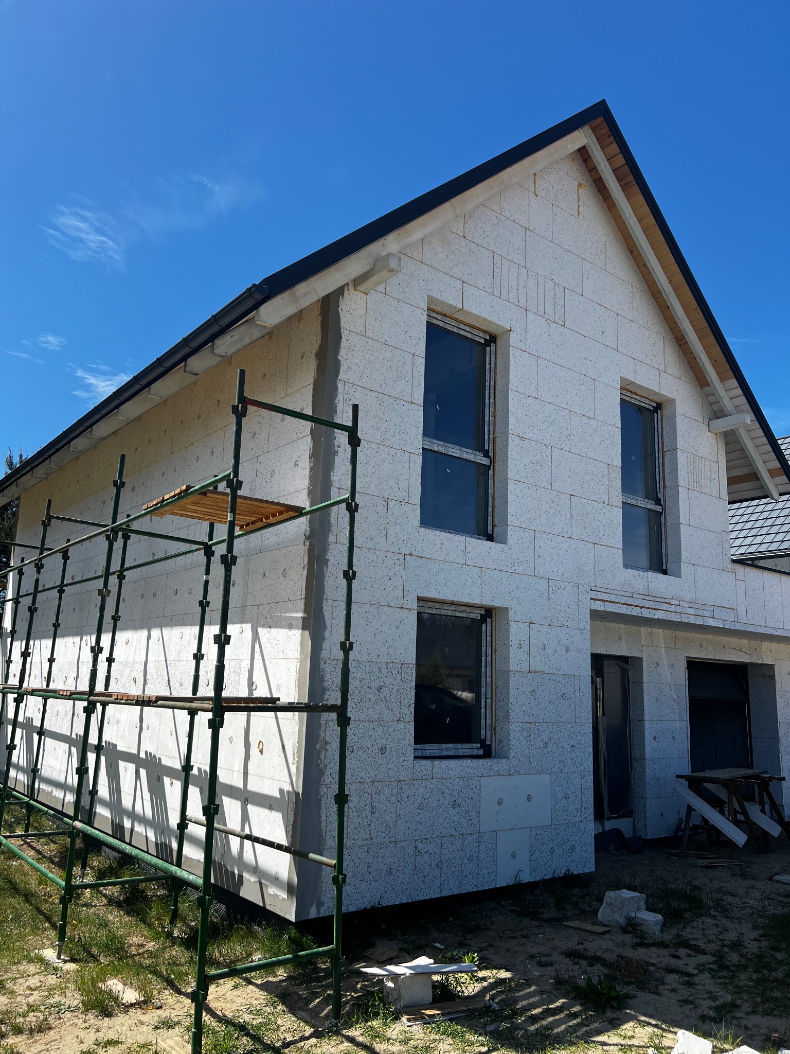 Budowa domu z białych bloków, widoczne rusztowanie i zamontowane okna z czarnymi ramami pod błękitnym niebem.