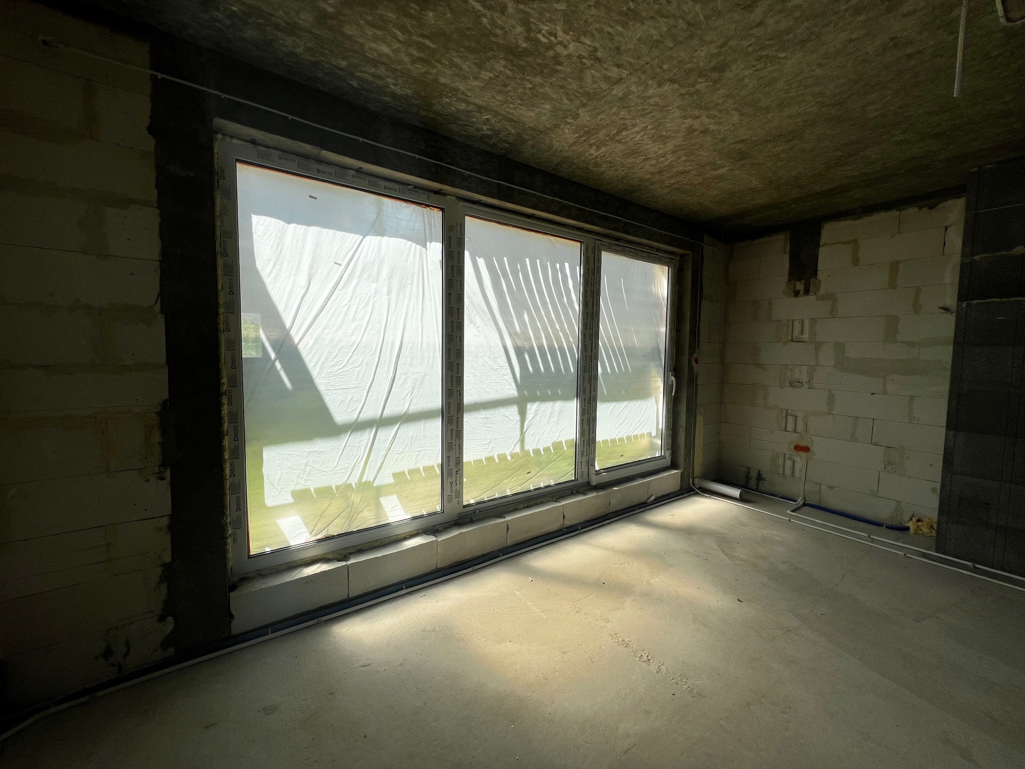 Trzyczęściowe okno, osłonięte folią ochronną, w surowym wnętrzu z betonową podłogą i ścianami z bloczków, widoczne instalacje.