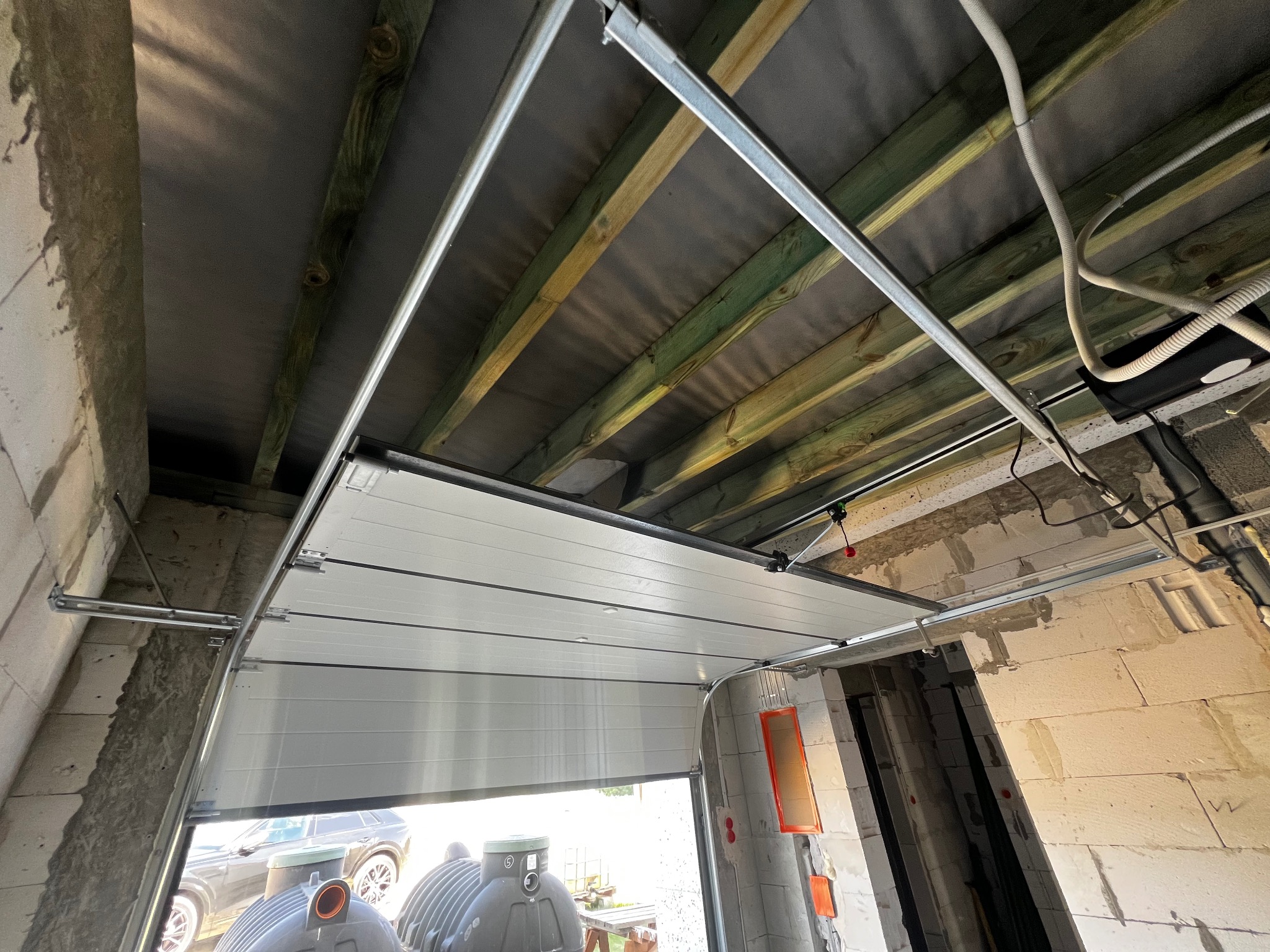 Montaż segmentowej bramy garażowej: widok na częściowo otwartą bramę z widocznym mechanizmem prowadnic i mocowań do sufitu garażu, w tle widoczne dwa zbiorniki na deszczówkę i fragment...