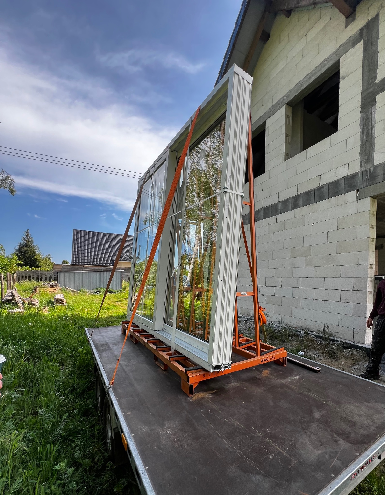 Nowe okno na przyczepie, zabezpieczone pomarańczowymi pasami, gotowe do montażu w surowym budynku z białych bloczków.