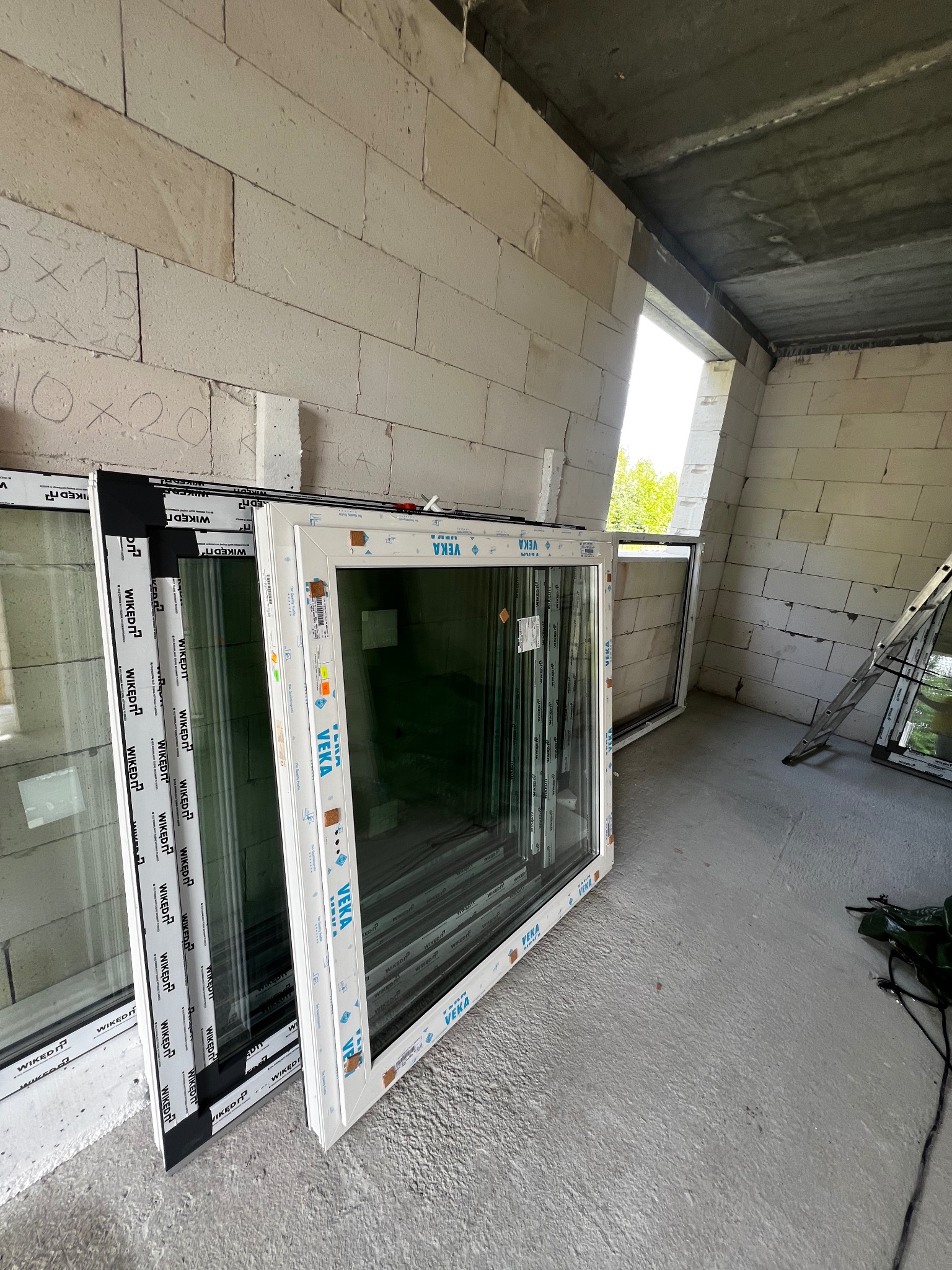 Nowe okna, białe i czarne ramy, oparte o surową ścianę z betonu komórkowego w trakcie budowy, widoczne oznaczenia producenta VEKA i WIKĘD.