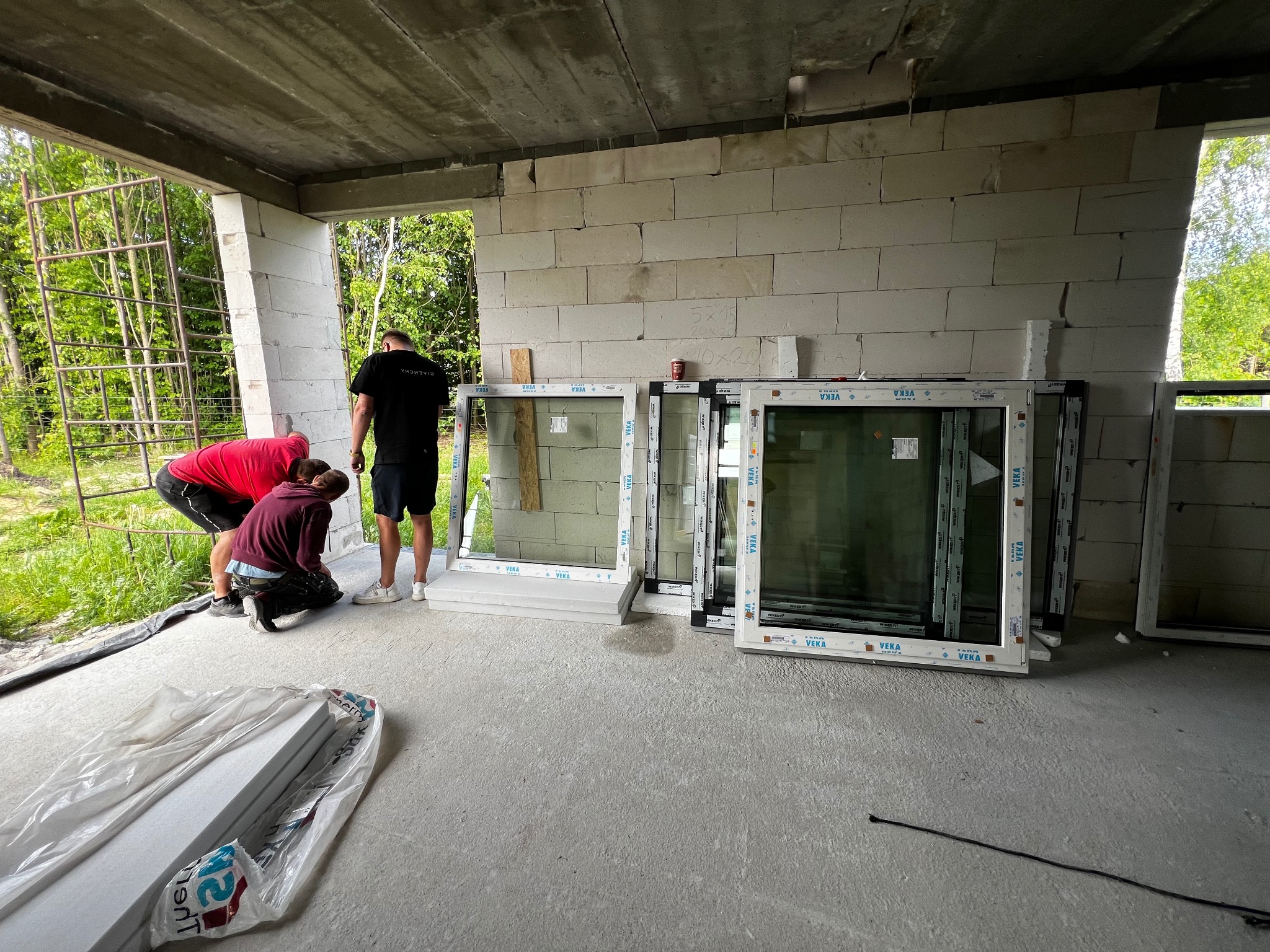 Trzech mężczyzn przygotowuje się do montażu białych okien z profili VEKA w surowym stanie budynku z betonu komórkowego; widoczne profile okienne w folii ochronnej.