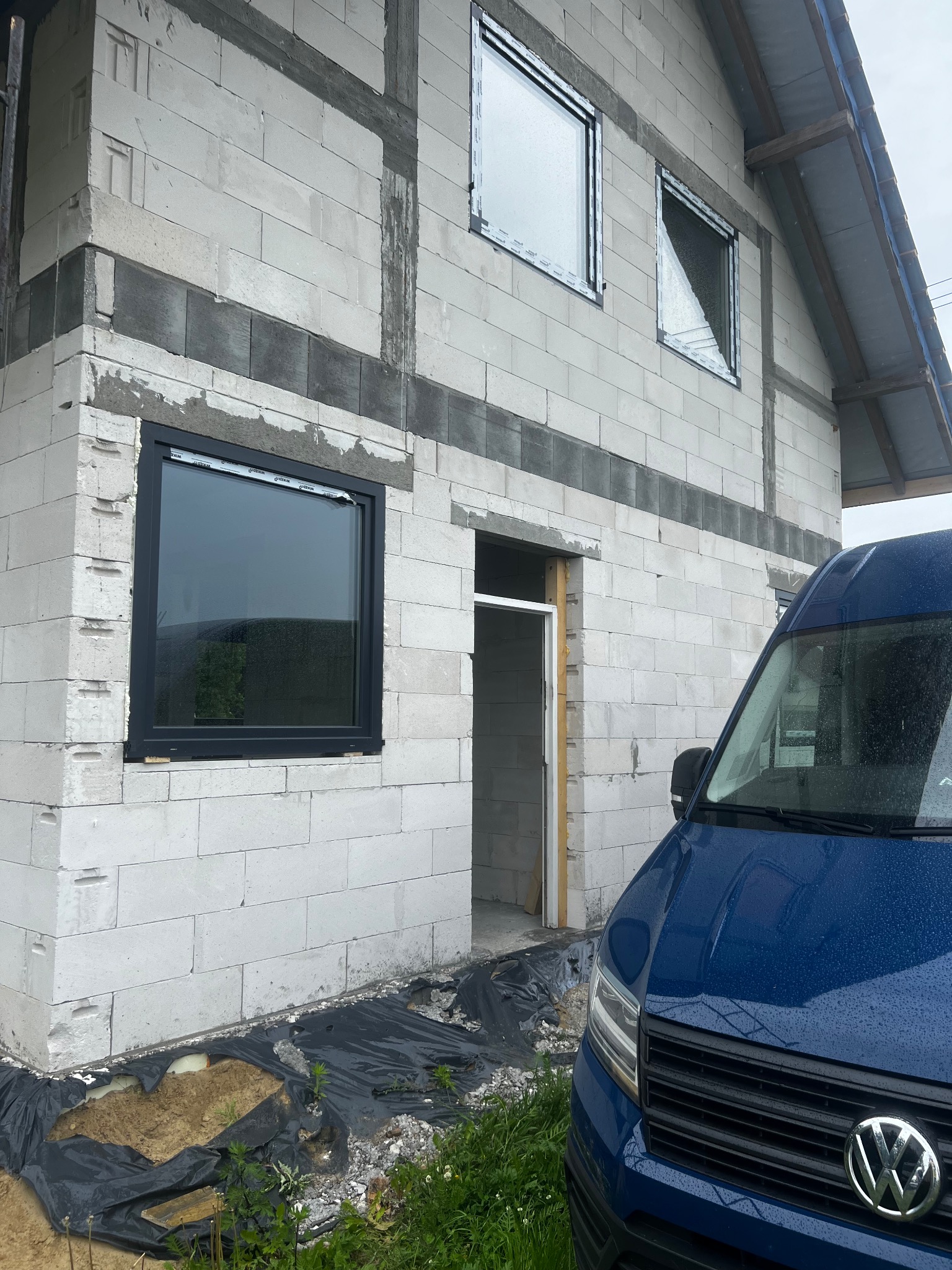 Surowy stan budynku z betonu komórkowego z zamontowanymi oknami w czarnych ramach, widoczny fragment niebieskiego samochodu dostawczego, w tle deszczowa pogoda.
