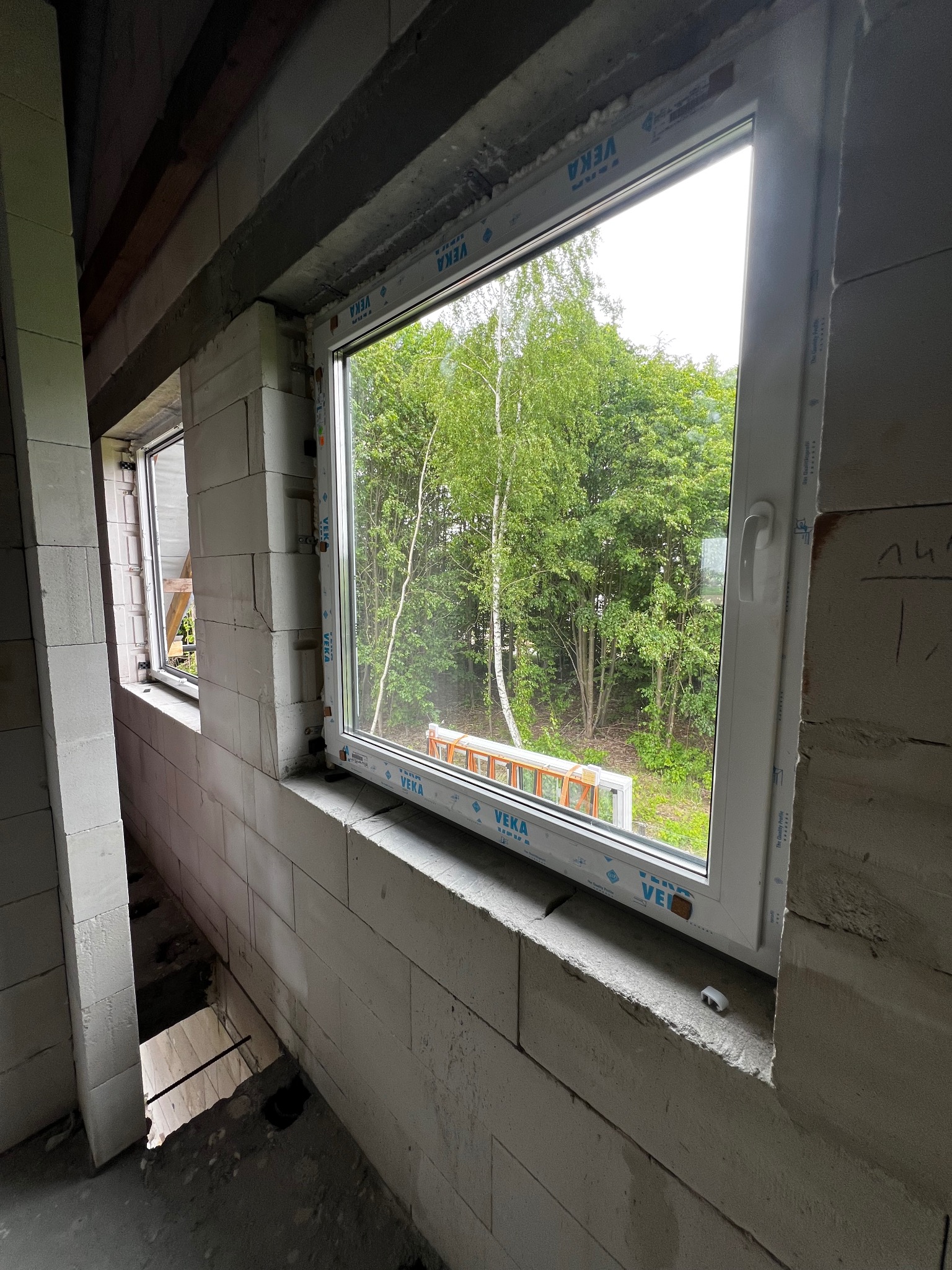 Nowo zamontowane białe okno z widokiem na zieleń drzew, w surowym stanie budowy, widoczne bloczki betonowe i otwór w podłodze.