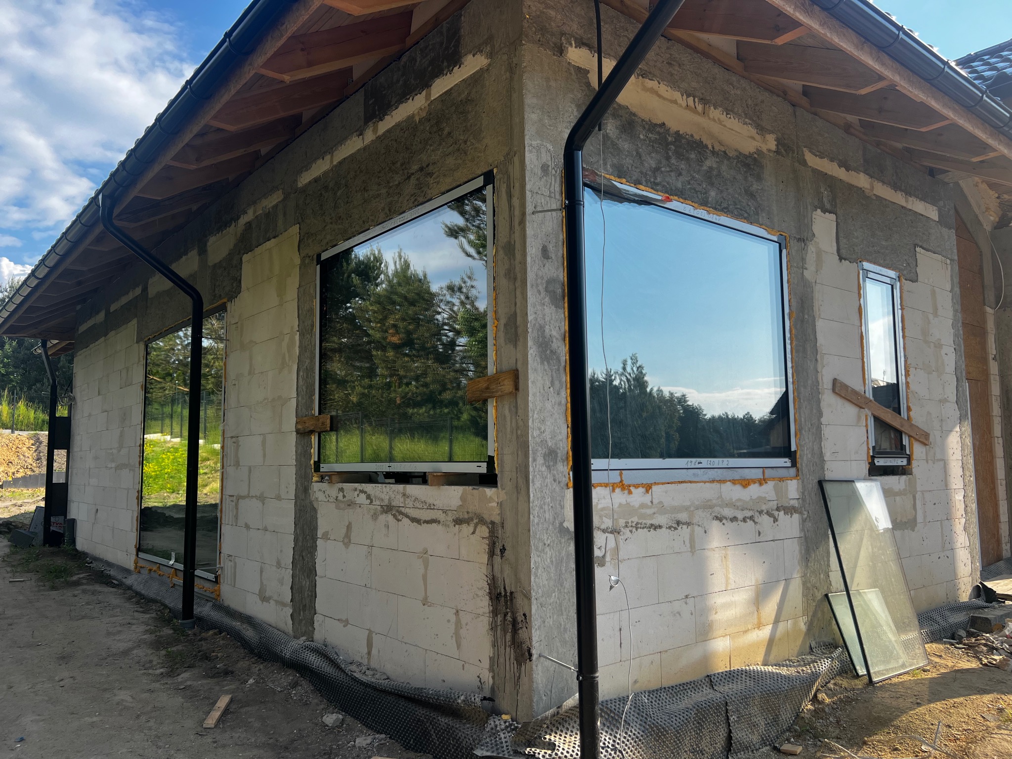 Surowy stan elewacji domu jednorodzinnego z zamontowanymi oknami z czarnymi ramami, widoczne odbicie drzew w szybach, przygotowane podłoże pod elewację.