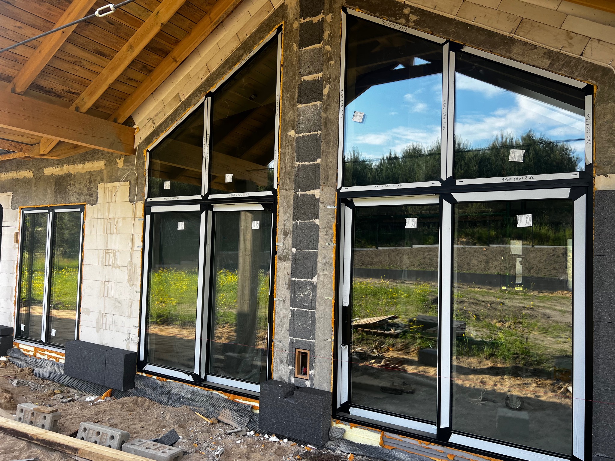 Montaż dużych, nowoczesnych okien z czarnymi ramami w surowym stanie budynku, widoczne niedokończone ocieplenie i elementy konstrukcyjne dachu.