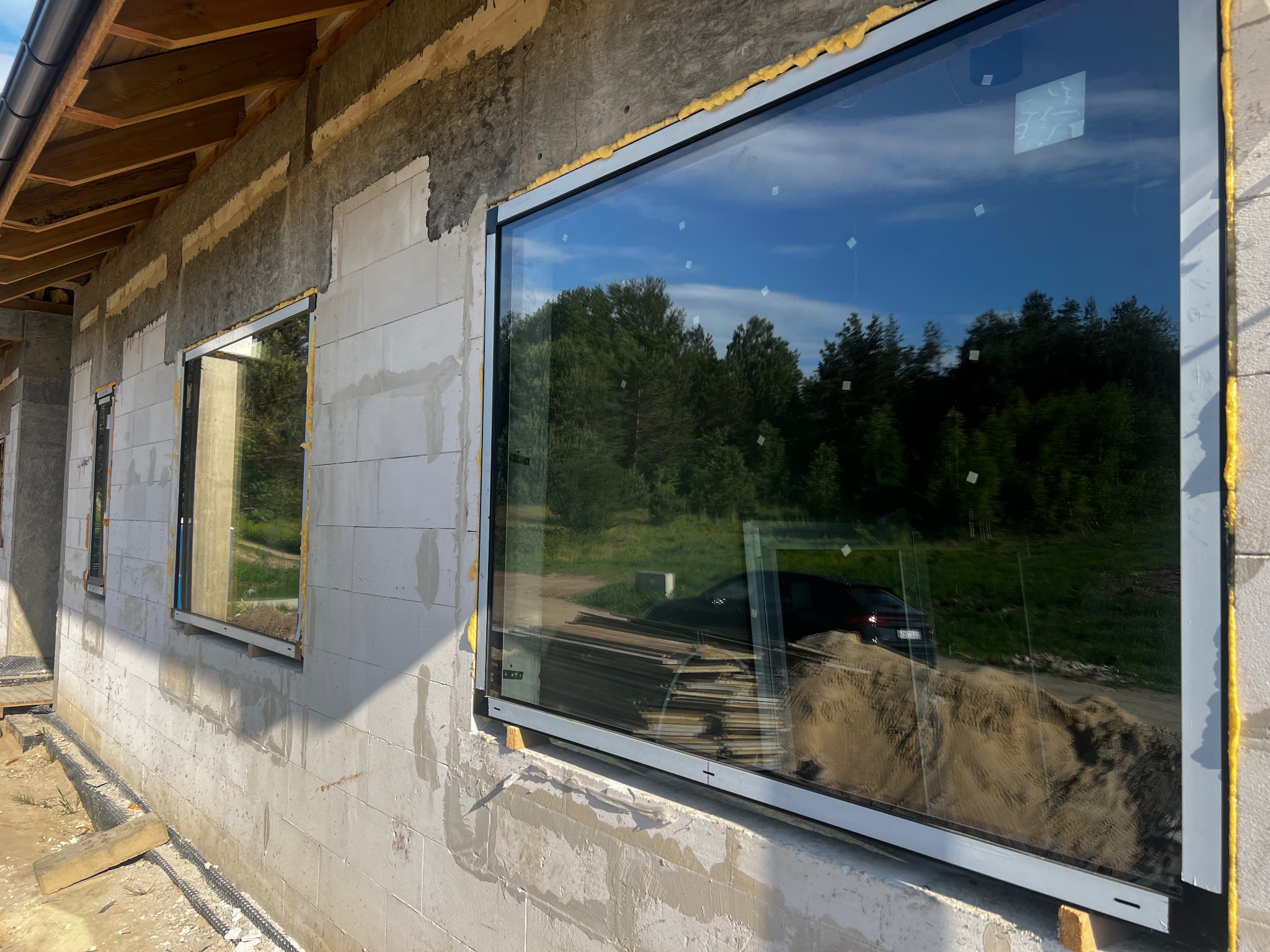 Montaż dużego, panoramicznego okna aluminiowego w surowym stanie budynku z betonu komórkowego, widoczne pianowanie uszczelniające i odbicie otoczenia w szybie.