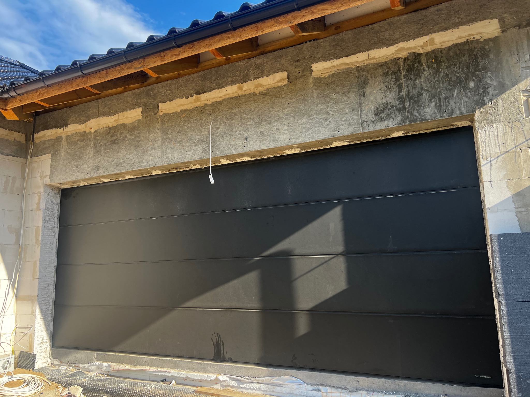 Ciemnoszara, segmentowa brama garażowa w trakcie montażu w surowym otworze budynku, widoczne materiały izolacyjne i instalacje elektryczne.