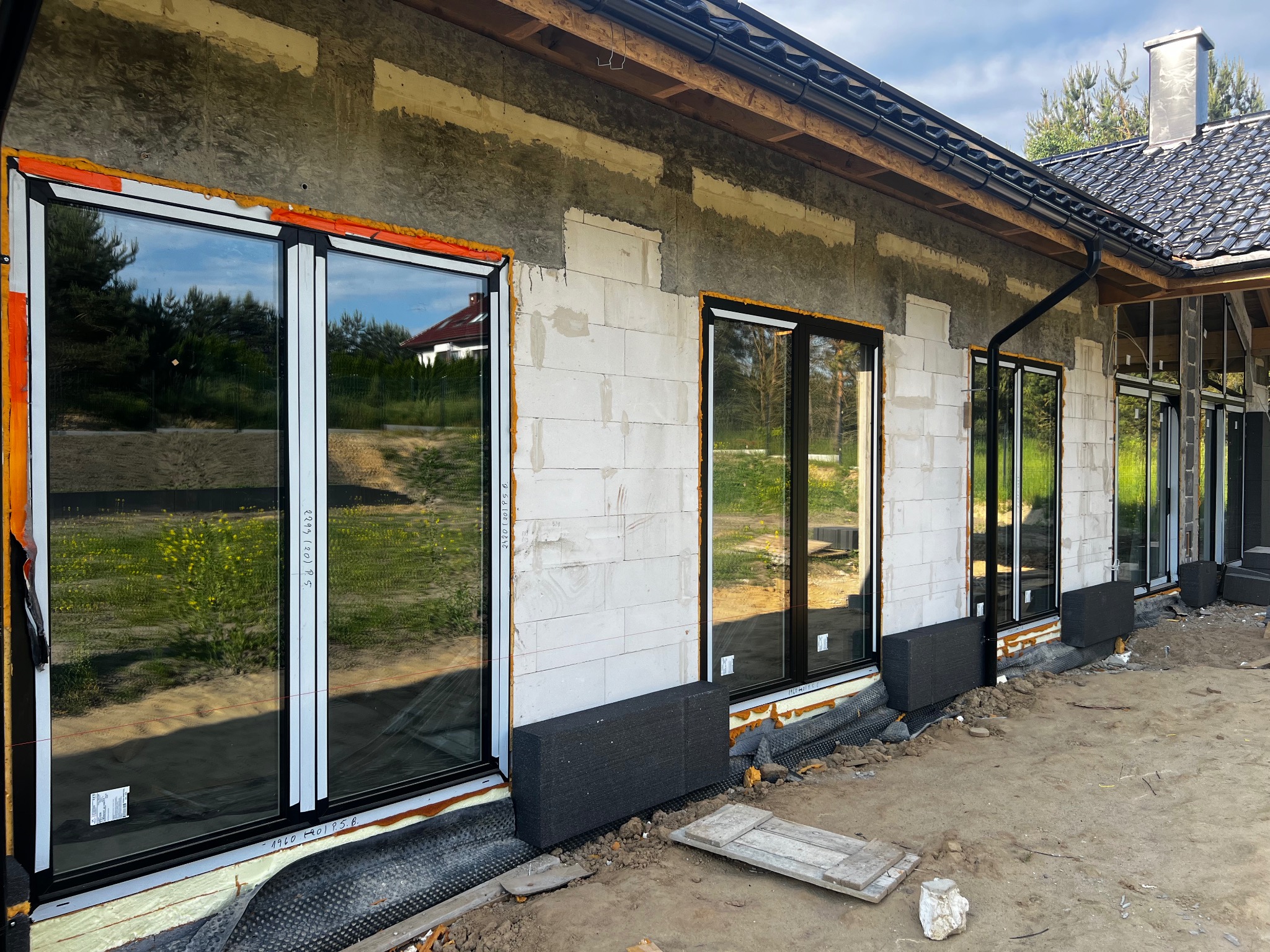 Montaż okien z czarnymi ramami w surowym stanie domu z betonu komórkowego, widoczne pianowanie i folia hydroizolacyjna przy fundamentach.