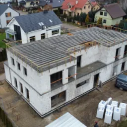 Dwukondygnacyjny budynek w stanie surowym otwartym z betonu komórkowego, widok z lotu ptaka, na dachu widoczna konstrukcja szalunkowa, obok zaparkowany bus firmy budowlanej i palety z materiałem...