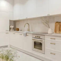 Jasna kuchnia z białymi szafkami, złotą armaturą i sprzętami AGD w stylu retro, z blatem wykończonym w jasnym odcieniu i dekoracyjnymi elementami takimi jak drewniane deski do krojenia i gałązki...