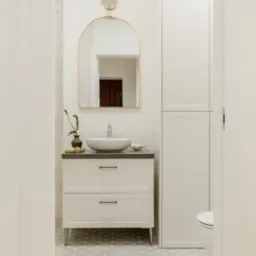 Jasna łazienka z umywalką nablatową, szafką z dwiema szufladami na nóżkach, lustrem w złotej ramie i złotym kinkietem. Na blacie umywalki dekoracje: roślina w wazonie i muszla.